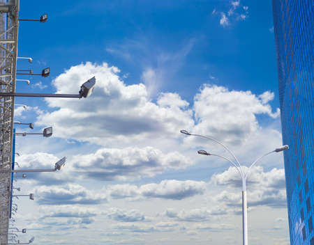 Many light fixtures for billboard backlighting, pole of a street lighting and fragment of the modern multistory office building against the background of sky with clouds in daytimeの写真素材