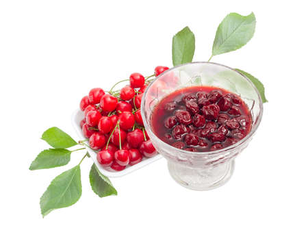 Cherry jam with whole fruits in the glass dessert stem bowl against of the fresh cherries with the stalks and leaves in the foam food container on a light backgroundの写真素材