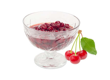 Cherry jam with whole fruits in the glass dessert stem bowl and three fresh cherries with the stalks and leaf beside on a light backgroundの写真素材