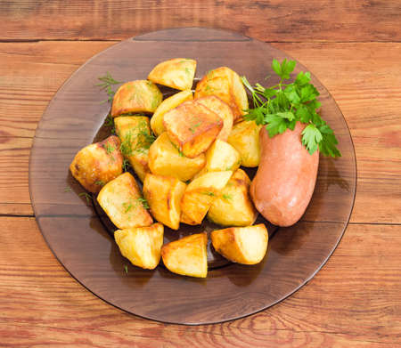 Top view of the dark glass dish with fried potatoes sprinkled by chopped dill, fried wieners and twig of parsley closeup on a surface of old wooden planksの写真素材