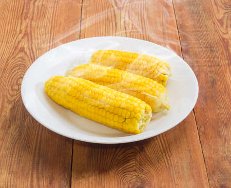 Three freshly boiled whole ears of sweet corn on a white dish on an old wooden surfaceの写真素材