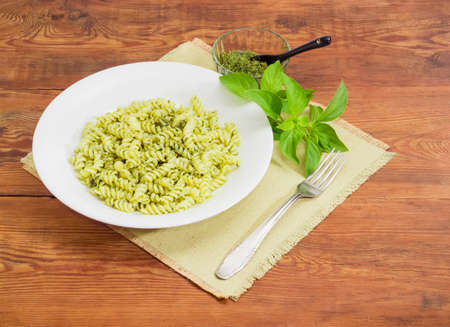 Cooked spiral pasta with sauce pesto on the white dish, pesto in small glass bowl with spoon, fork and basil twig on a napkin on a surface of old wooden planksの写真素材