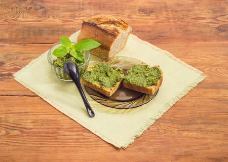 Two sandwiches with sauce pesto, sauce pesto in small glass bowl with small black spoon and decorated with green basil twig, part of bread on a napkin on a surface of  old wooden planksの写真素材
