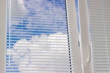 Fragment of the modern plastic window with Venetian blinds and view of the sky with clouds across slats of a window blindの写真素材