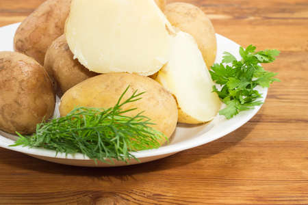 Fragment of white dish with potatoes boiled in their jackets decorated with twigs of parsley and dill on a surface of old wooden planks closeupの写真素材