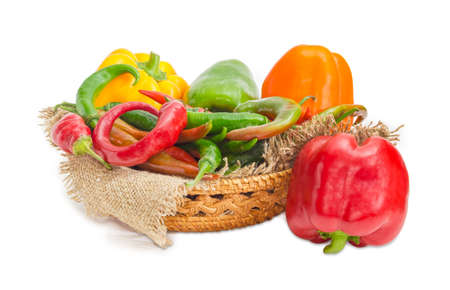 Red and green chili and several multi-colored bell peppers on a sackcloth in small wicker basket and beside on a white backgroundの写真素材