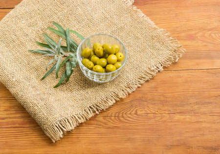 Canned green olives in small glass bowl and olive branch on a sackcloth on a surface of an old wooden planksの写真素材