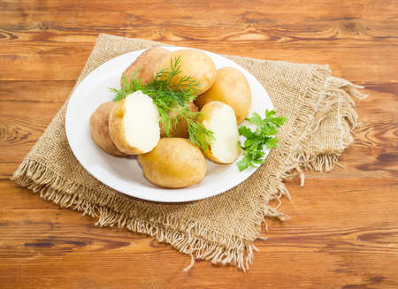 Two halves and several whole potatoes boiled in their jackets decorated with twigs of parsley and dill on the white dish on a surface of old wooden planks with sackclothの素材 [FY31087672232]