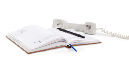 Open business diary and black pen on it on background of the white handset of a landline telephone lying down beside at shallow depth of field on a white backgroundの写真素材