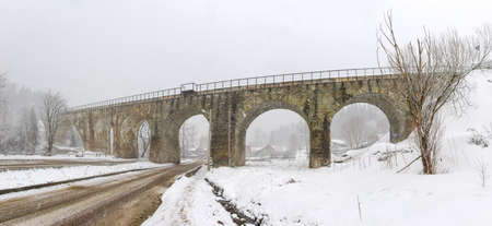 Old railway viaduct made of stone masonry over valley with highway in Carpathian Mountains during heavy snowfallの写真素材