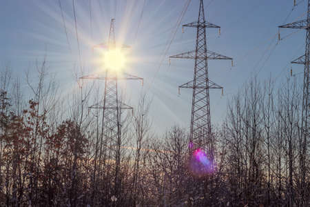 Several overhead power lines with steel lattice structures towers over small trees on a background...