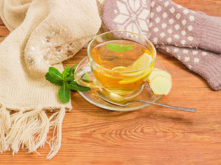 Glass cup of tea with lemon and ginger root slices, mint leaf on glass saucer with spoon closeup on an old wooden surface on background of the women's woolen mittens and knitted scarf の写真素材