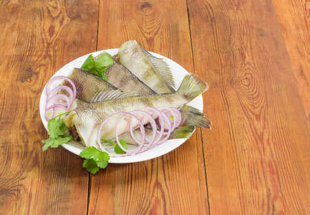 Several uncooked carcasses of the notothenia fish without of the heads, cilantro twigs and round slices of red onion on a white dish on an old rustic wooden tableの写真素材