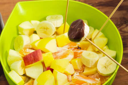 Fragment of chocolate fondue - piece of fruit on a bamboo wooden skewer, dipped in liquid chocolate over container with pieces of different fruits closeup at shallow depth of field on a rustic tableの写真素材