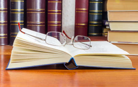 Classic mens eyeglasses lying on the open book in blue hardcover at selective focus with blurred  book text on a wooden table against of the other booksの写真素材