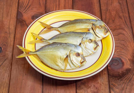 Three smoked Selene setapinnis, also known as Atlantic moonfish or Vomer setapinnis, on the white and yellow dish on a surface of dark-colored wooden planksの写真素材