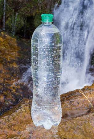 Plastic bottle of drinking water standing on the wet stone and covered with drops of water at selective focus on a blurred background of the mountain stream with small waterfallの写真素材