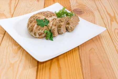Partly sliced baked chopped pork liver pate decorated with dill and parsley twigs on a white square dish closeup at selective focus on a light colored wooden surfaceの写真素材