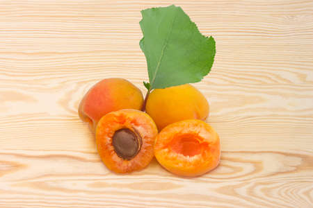 Two whole ripe apricots and one cut in half with leaf on a light colored wooden surface