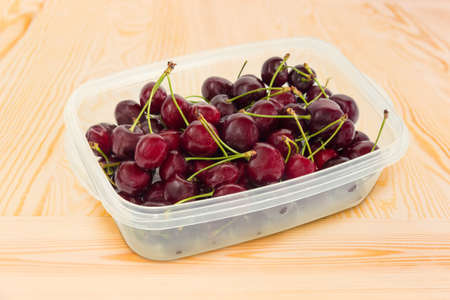 Dark red sweet cherries with stalks in plastic container at selective focus on a light colored wooden surfaceの写真素材