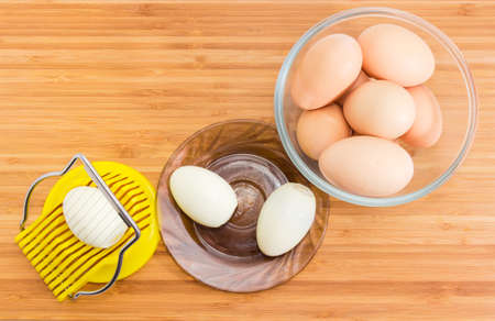 Top view of egg slicer with egg in it, two boiled peeled eggs on saucer and not cleaned eggs in a glass bowl on a wooden bamboo surfaceの写真素材