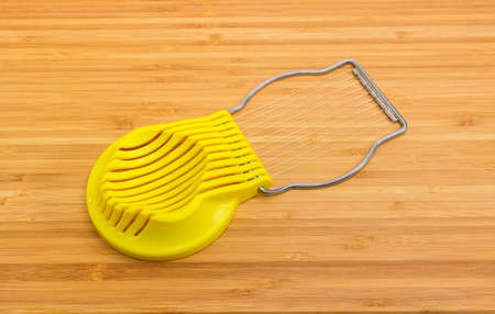 Manual egg slicer with yellow plastic slotted dish and open steel plate of wires on a bamboo cutting boardの写真素材