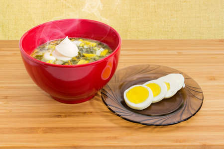 Hot sorrel soup also known as green borscht with chopped egg and sour cream in red bowl and sliced boiled egg beside to it on a wooden tableの写真素材