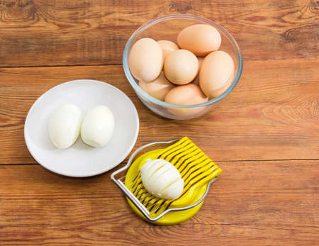 Egg slicer with partly sliced egg in it, two boiled peeled eggs on a saucer and not cleaned eggs in a glass bowl on an old rustic tableの写真素材