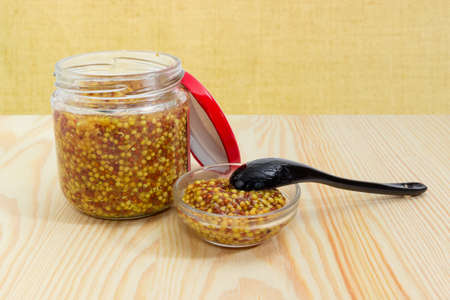 Coarse whole-grain French mustard made from black and yellow mustard seeds in glass jar with open red lid and small glass bowl with black spoon on a light colored wooden surfaceの写真素材