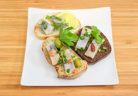 Three different open sandwiches made of slices of fillet of pickled Atlantic herring and various bread on the square dish on a wooden bamboo surface の写真素材