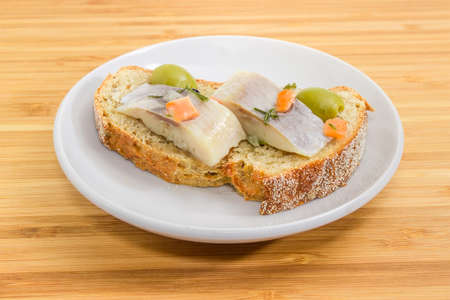 Open sandwich made of brown sprouted bread, slices of fillet of pickled Atlantic herring and green olives on a saucer closeup at selective focus on a wooden bamboo surfaceの写真素材
