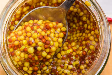 Top view of the whole-grain French mustard made from black and yellow mustard seeds on tea spoon in small glass jar closeupの写真素材