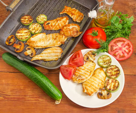 Top view of grilled chicken meat, vegetables and mushrooms on the white dish and grill pan, raw tomatoes cucumber and greens on an old wooden rustic table
の写真素材