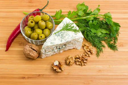 Piece of the blue cheese among of green olives in small bowl, pod of chili, greens and walnuts closeup on the bamboo cutting board 
の写真素材