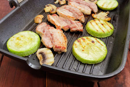 Grilled slices of streaky bacon with vegetable marrows and mushrooms on the grill pan closeup at shallow depth of field on a dark wooden rustic tableの写真素材