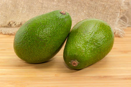 Two whole green-skinned pear-shaped avocado fruit on a wooden bamboo surface