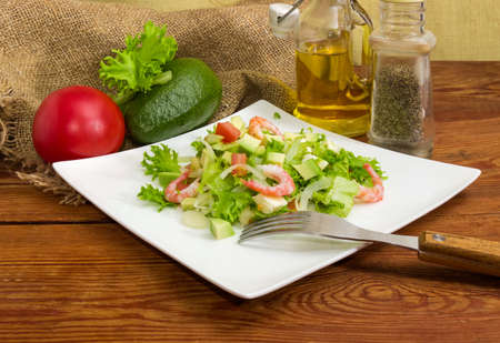 Salad made of sliced avocado, vegetables, diced mozzarella cheese and decorated with shrimps tails on square dish, fork and some ingredients for its preparationの写真素材