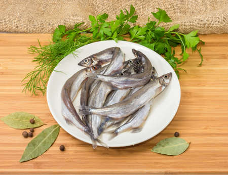 Defrosted uncooked capelins on the white dish, bay leaf, allspice and fresh parsley and dill on a wooden surfaceの写真素材