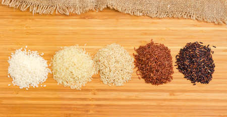 Small piles of raw short-grained white and parboiled rice, long-grained brown, red and black rice on a wooden bamboo cutting boardの写真素材