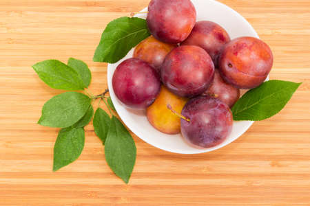 Top view of the white bowl with ripe purple plums and leaves of plum tree close-up on a bamboo wooden surfaceの写真素材