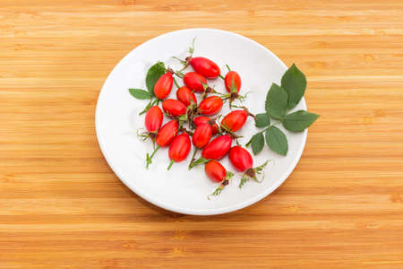 Fresh red rose hips with leaves on a white saucer on a bamboo wooden surfaceの写真素材