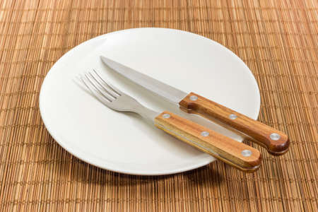 Fork and knife with wooden handles on empty round white dish on the bamboo table mat close-up at...