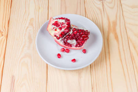 Piece of splitted ripe pomegranate fruit and several fallen out seeds on the white saucer on a wooden rustic tableの写真素材