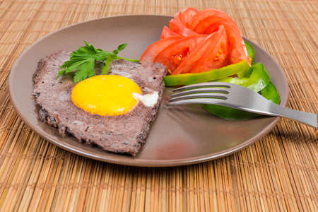 Fried egg sunny side up prepared by beating egg white with black olives and unbroken yolk, slices vegetables on brown dishの写真素材