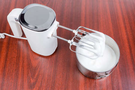 White electric hand-held mixer with two beaters and beaten eggs in stainless steel pot  on a wooden tableの写真素材