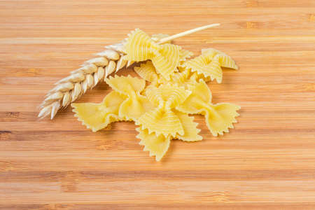 Several uncooked bow-tie shape pasta also known as farfalle pasta and ear of wheat close-up on a wooden surfaceの写真素材