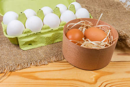 Brown chicken eggs in round wooden box on background of white eggs in open carton made of recycled...