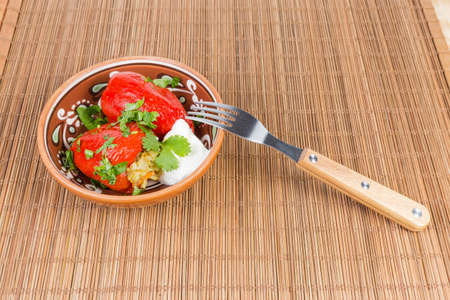 Two cooked stuffed red bell peppers with chopped greens in ceramic bowl and fork on the bamboo table matの写真素材