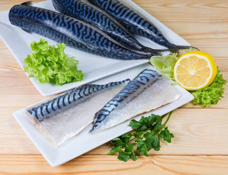 Salted fillet of Atlantic mackerel in vegetable oil on dish against of raw headless fish carcasses, lemon and greens on a rustic tableの写真素材