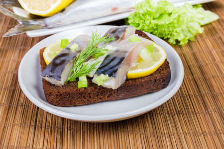 Open sandwich with slices of salted Atlantic mackerel, dill and lemon on brown bread, sprinkled with chopped green onion on saucer on a bamboo table mat close-up at selective focusの写真素材